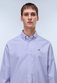 Light blue striped button-up shirt with a collar and white buttons. Features a small flag logo on the left chest. Soft cotton material.