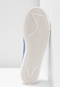 Baskets en daim bleu montrant une semelle en caoutchouc blanche avec un motif en chevrons et des détails texturés. Positionnées à la verticale sur un fond neutre.