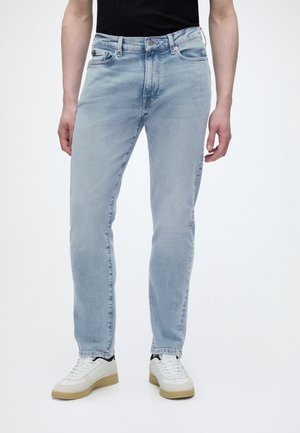 Homme portant un jean slim bleu clair et des baskets blanches, debout devant un fond blanc uni.