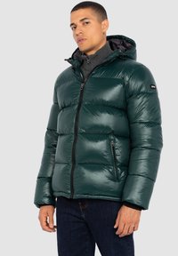 Chaqueta de plumas verde con textura brillante, cierre de cremallera, dos bolsillos laterales y capucha. Se lleva sobre un suéter de punto oscuro y unos vaqueros.
