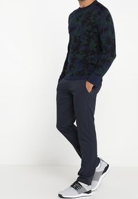 Un homme portant une chemise à manches longues à motif camouflage sombre, un pantalon bleu marine et des baskets grises, se tenant avec les mains dans les poches contre un fond blanc.