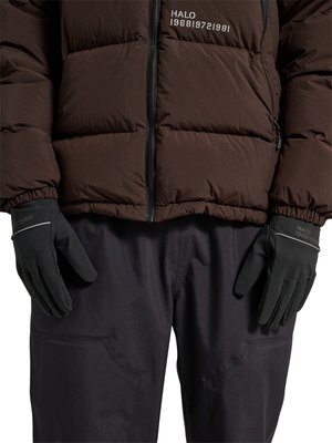 Osoba nosząca brązową kurtkę puchową i czarne rękawiczki, obie z napisem "HALO 1968|972|981", oraz czarne spodnie z przednimi kieszeniami.
