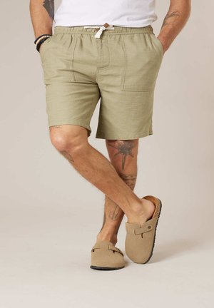 Homme portant un short beige avec des poches avant, un cordon blanc, des sandales beiges à enfiler, et des tatouages visibles sur les jambes, se tenant jambes croisées.