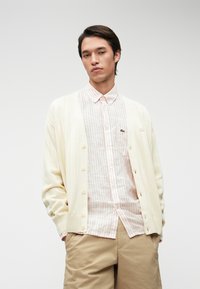 Κρέμα ζακέτα με κάθετη ριγωτή υφή, με κουμπιά μπροστά; κάτω από αυτήν, ροζ ριγέ πουκάμισο; χακί βερμούδα. Χωρίς ορατά σχέδια ή λεπτομέρειες.