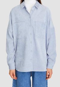 Jasnoniebieska, oversize koszula wykonana z miękkiego materiału, z zapięciem na guziki z przodu, dwoma kieszeniami na piersi oraz długimi rękawami o luźnym kroju.