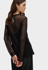 Svart genomskinlig blus med prickmönster, långa ärmar och en nyckelhålsdetalj bak. Texturerad tyg med lätt genomskinlighet.