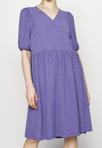 Robe violette avec manches courtes bouffantes, encolure en V et pois blancs. Design évasé avec taille froncée et ourlet à hauteur des genoux.