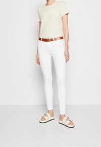 Camiseta ligera color beige con logo, jeans skinny blancos, cinturón marrón y sandalias color crema con acentos metálicos, mostrando un atuendo casual.