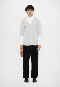 Camicia a righe bianche e grigie con colletto, abbinata a pantaloni neri a gamba larga. Il modello tiene in mano una piccola custodia marrone e indossa scarpe nere.