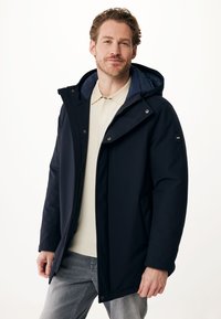 Veste imperméable bleu marine foncé avec capuche, présentant une texture lisse, une fermeture éclair, des accessoires en métal argenté et une étiquette de marque sur la manche.