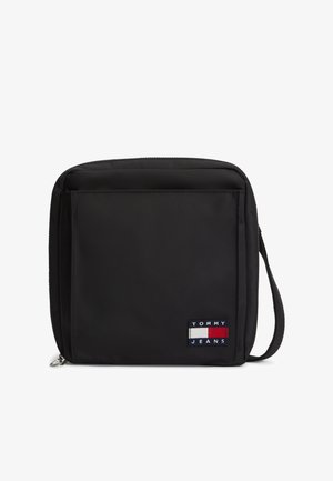 Czarna kwadratowa torba na ramię z przednią kieszenią, zamknięciem na suwak, paskiem na nadgarstek oraz naszywką z logo Tommy Jeans w kolorach czerwonym, białym i granatowym.