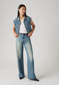 Leren spencer in lichtblauw met een kraag en mouwloos ontwerp. Gepaard met hoge taille, wijde lichtblauwe jeans, die een zachte vervaging tonen.