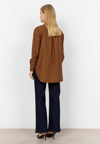 Brown button-up shirt with long sleeves, side slits, and a curved hem, paired with dark flared jeans and black heeled sandals.