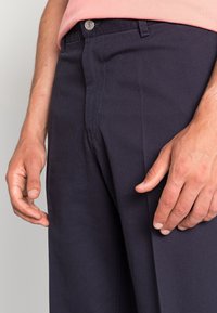 Marineblaue Hose mit geradem Schnitt, ausgestattet mit einer Vordertasche, Knopfverschluss und leichter Textur. Hand in der Nähe der Taschennaht positioniert.
