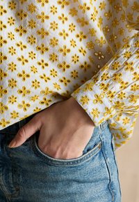 Bluză albă cu flori galbene brodate, având un manșon adunat și un buzunar vizibil din blugi albaștri.