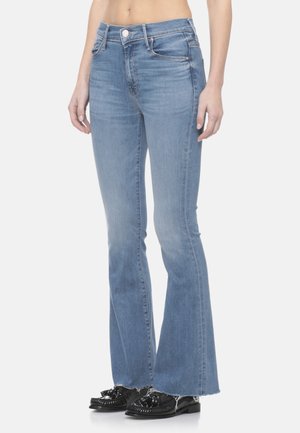 Jeans aderenti in denim blu con leggera svasatura e orlo grezzo indossati da una persona in piedi, abbinati a mocassini neri con nappine.