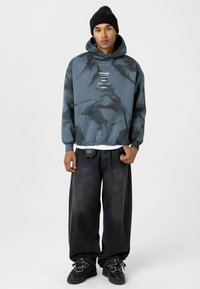 Pullover hoodie in blauw-grijs tie-dye met witte grafische print, voorzak en hoodie met trekkoord. Gecombineerd met wijde zwarte spijkerbroek en zwarte sneakers.