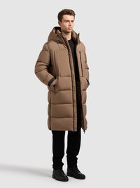 Bruine pufferjas met capuchon, uitgevoerd met een ritssluiting, zijzakken en een doorgestikt ontwerp. Gecombineerd met zwarte broek en schoeisel.