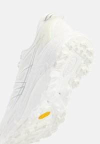 Hvit atletisk sko med teksturert overdel, forsterket hæl og en robust gummisåle med markante knotter og en gul Vibram-logo.