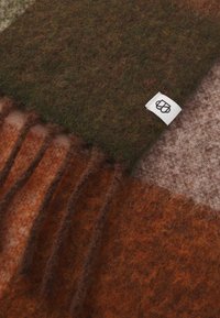 Écharpe en feutre avec un motif à carreaux marron, vert et orange, présentant des bords frangés et une petite étiquette de marque blanche. Texture douce.