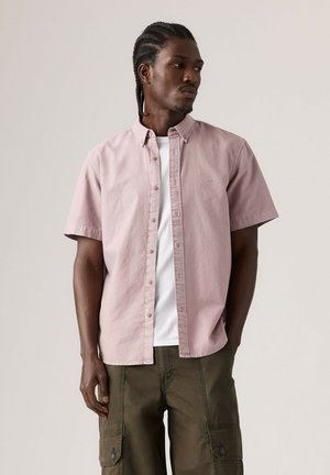 Homme aux cheveux tressés portant une chemise rose pâle à manches courtes boutonnée sur un t-shirt blanc et un pantalon cargo olive, regardant sur le côté.