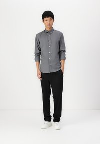 Camisa gris de manga larga con botones y cuello, combinada con pantalones negros y zapatillas blancas. La camisa tiene las mangas arremangadas y un corte ajustado.