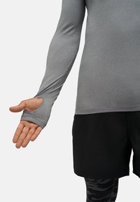 Szara sportowa koszulka z długim rękawem z otworami na kciuki, w zestawieniu z czarnymi szortami. Gładki materiał z dopasowanym krój i minimalną ilością szwów.