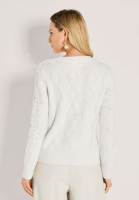 Frau mit blonden Haaren, die einen weißen strukturierten Strickpulli mit Rautenmuster und cremefarbenen Hosen trägt, von hinten gezeigt.