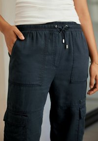 Pantaloni cargo blu navy con vita elastica, chiusura a lacci, tasche laterali e tasche applicate sulle cosce. Tessuto liscio.