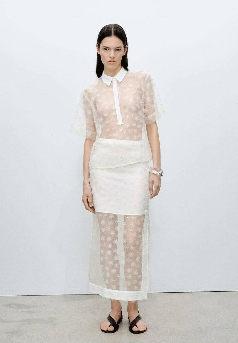 Blusa trasparente con maniche corte, colletto bianco, indossata sopra una gonna abbinata; motivo floreale; indossata con sandali neri.