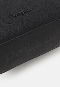 Czarna teksturowana saszetka z wytłoczonymi okrągłymi wzorami i nazwą marki "Desigual". Wykonana z materiału imitującego skórę o gładkim wykończeniu.