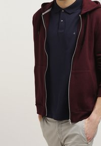 Mężczyzna w bordowym rozpinanym hoodie, z granatową koszulą polo i beżowymi spodniami, stoi z jedną ręką w kieszeni.