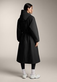 Egy hosszú fekete kapucnis kabátot, fekete leggingset, fehér zoknit és fehér tornacipőt viselő nő oldalra áll egy sima háttér előtt.