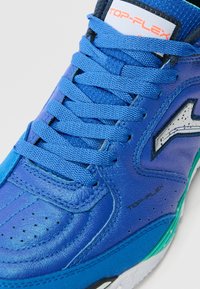 Scarpa atletica blu con tomaia testurizzata, lacci, colletto imbottito e dettagli verdi. Presenta un logo e sezioni perforate per la ventilazione.