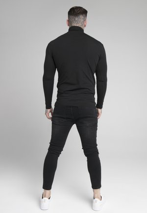 Czarna, długa koszulka z golfem w parze z czarnymi, dopasowanymi dżinsami i białymi trampkami, pokazana od tyłu na jasnoszarym tle.