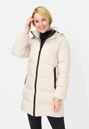 Lächelnde Frau mit kurzen blonden Haaren, die einen hellbeigen Steppmantel mit schwarzem Reißverschluss und Taschen trägt und vor weißem Hintergrund posiert.