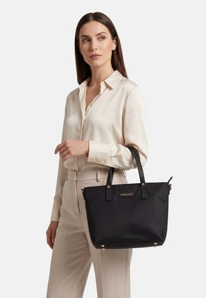 Žena v béžové blůze a kalhotách drží černou tašku Vivisence s koženými uchy a zlatými kovovými prvky.