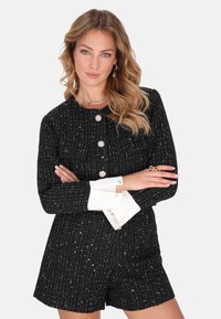 Romper negro de textura, de manga corta con detalles en plateado, que presenta botones de perla y puños blancos. Diseño clásico con un ajuste estructurado.