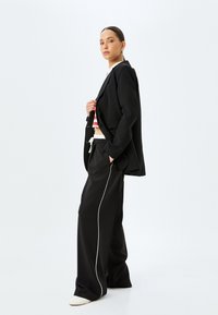 Sort blazer og brede sorte bukser med hvid piping. Modellen har en stribet top på under, som fremhæver lagdelte teksturer.