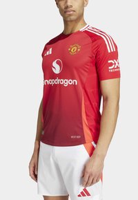 Maillot rouge de Manchester United à manches courtes, avec des bandes blanches, un logo et des marquages. Le tissu est texturé et évacue l'humidité.