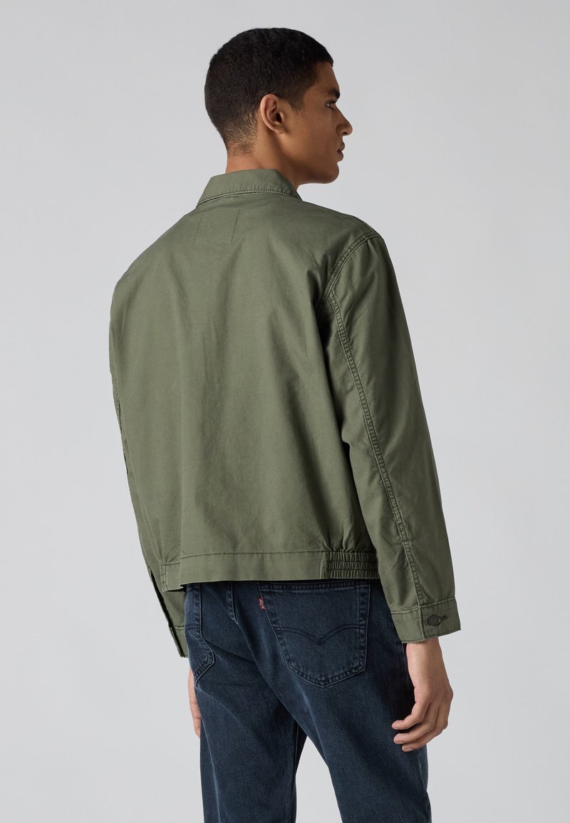 Hombre de pie de espaldas, vistiendo una chaqueta verde oliva y unos vaqueros azul oscuro sobre un fondo gris liso.