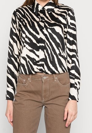 Czarnobiała koszula w paski zebry z kołnierzykiem, długimi rękawami i błyszczącą fakturą; w połączeniu z jasnobrązowymi spodniami.