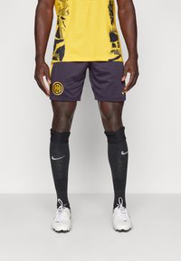 Geel sportshirt met zwarte patronen, marineblauwe shorts met een logo, zwarte sokken met een logo en witte voetbalschoenen.