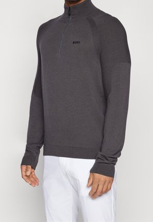 Mann trägt dunkelgrauen Viertel-Reißverschluss-Pullover mit gerippten Schultern und BOSS-Logo, kombiniert mit weißen Hosen vor einem einfarbigen hellen Hintergrund.
