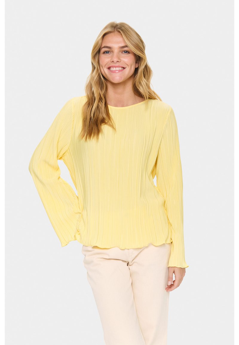 Donna sorridente con lunghi capelli biondi, che indossa una blusa lunga a maniche pleat gialla e pantaloni beige chiaro, in piedi davanti a uno sfondo semplice.