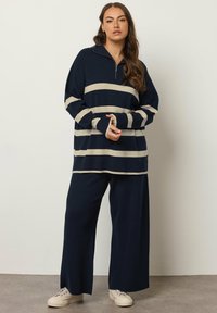 Maglione a righe blu navy e crema con design a mezza zip, vestibilità ampia e maniche lunghe, abbinato a pantaloni a gamba larga blu navy e sneaker beige.