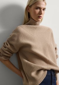 Maglione a costine beige con colletto alto, caratterizzato da motivi diagonali e maniche arrotolate, abbinato a jeans scuri.