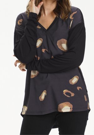 Mujer con una camiseta de manga larga negra con manchas abstractas en tonos marrón y beige, posando con un brazo cruzado sobre el cuerpo y la otra mano cerca del rostro.