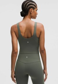 Kvind med fletninger, iført matchende olivengrøn ærmeløs crop top og højtaljede leggings, set bagfra mod en ensfarvet baggrund.