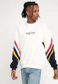 Hvit hettegenser med flerfargede stripete ermer (rød, blå, gul og marineblå) med en svart logo "KAOTIKO" på forsiden. Dongeribukser.